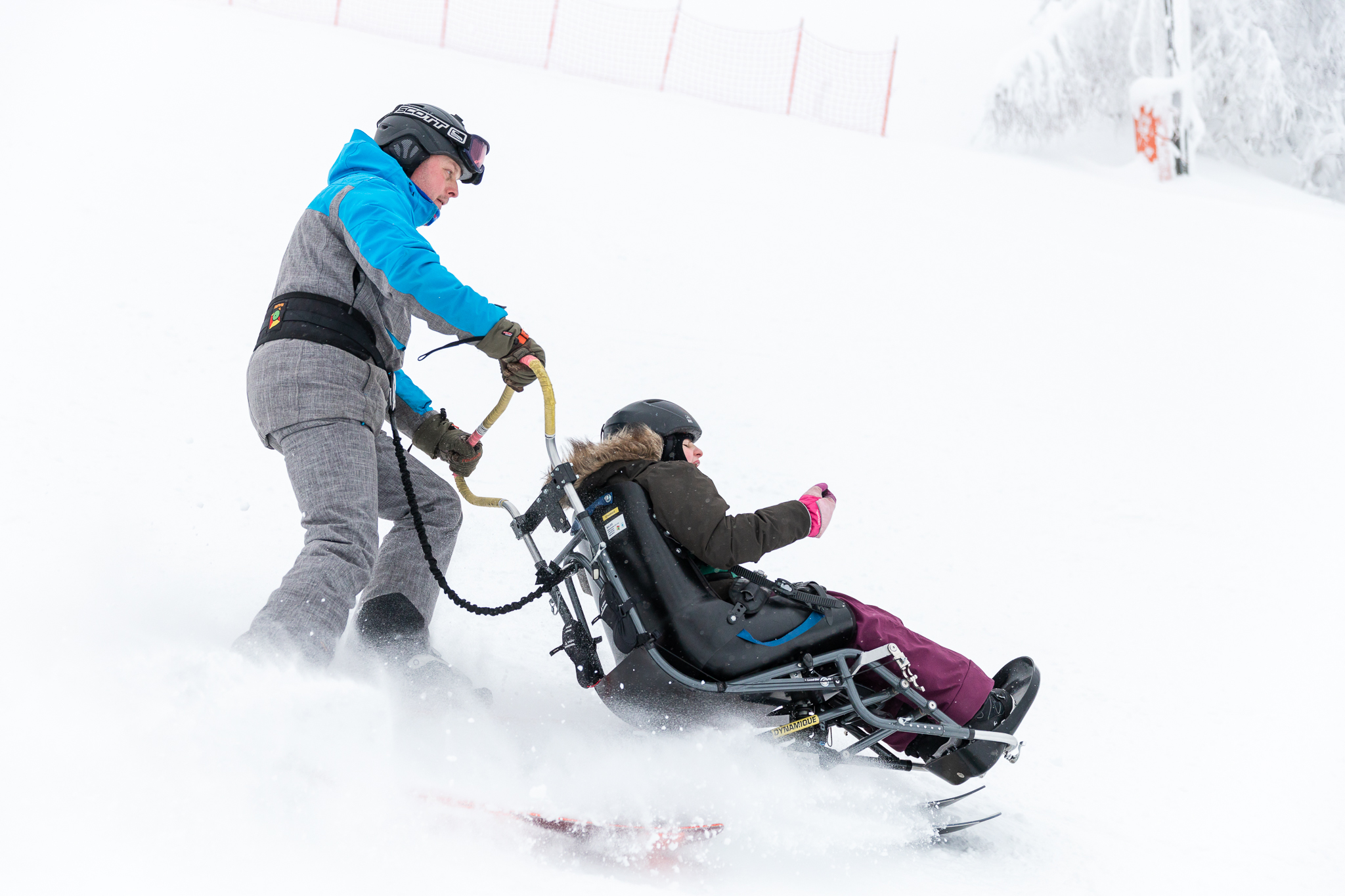 Kuvassa lapsi laskee avustajan kanssa laskettelukelkalla. Lumi pöllyää.
